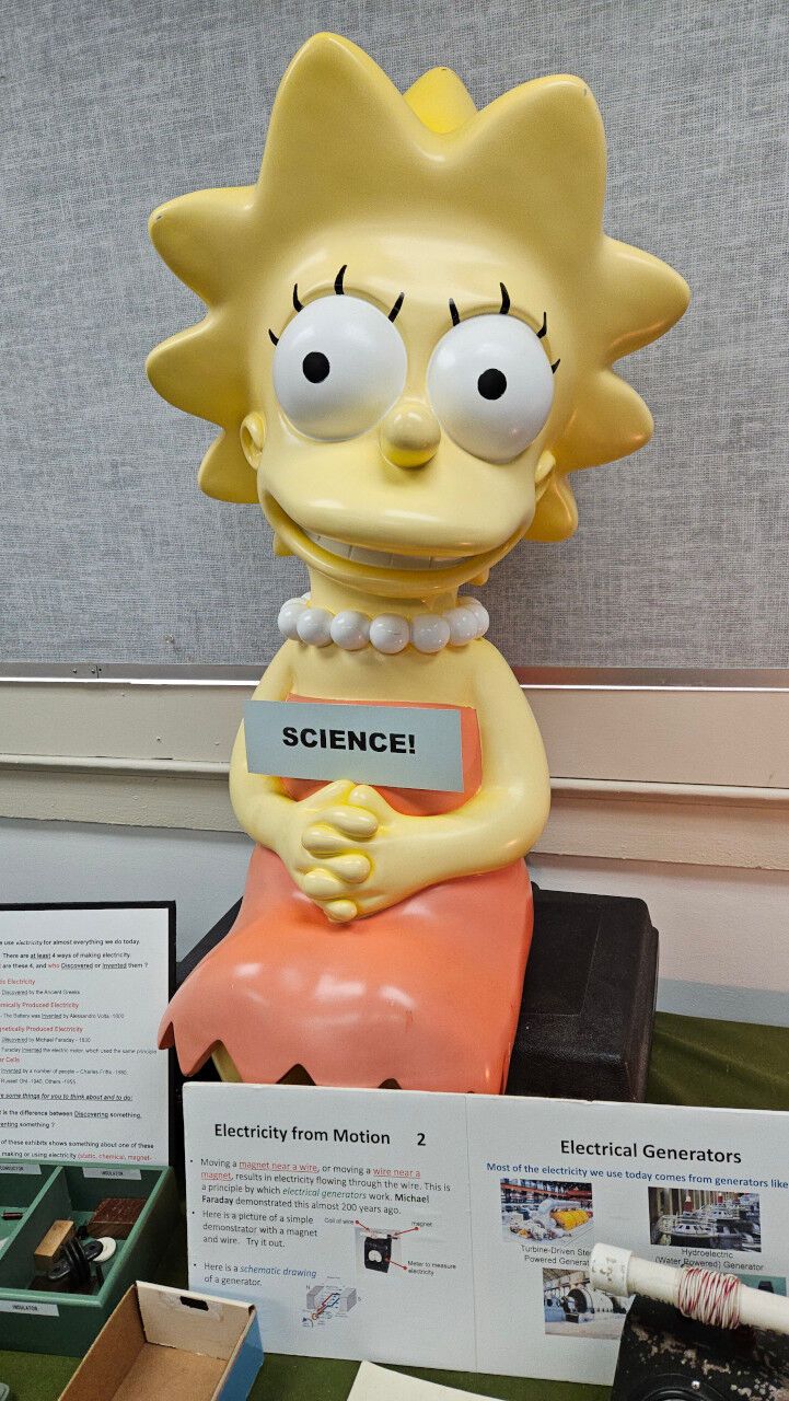 A large statue of Lisa Simpson, face on, with a sign saying SCIENCE propped up on her hands. Below are educational materials about electromagnetism.