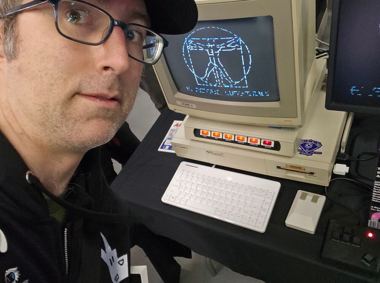 A middle aged man standing in front of an Amiga 1000. A demoscene demo titled Electric Lifeforms is playing on the screen.