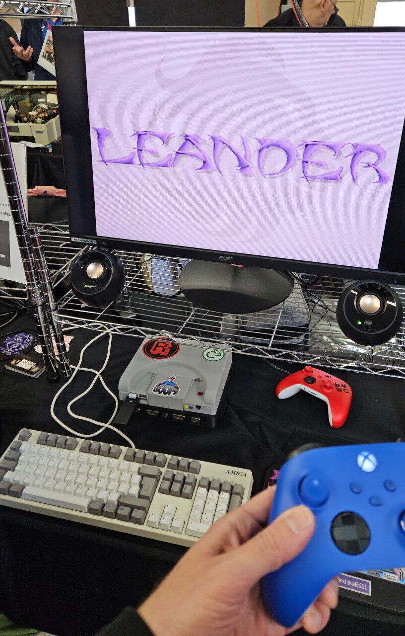 A monitor showing the intro screen for the game Leander. The monitor is hooked up to a game console. A person is holding a gamepad up.